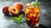 Refreshing peach iced tea with mint garnish in glass, served with fresh peach slices on dark stone background.