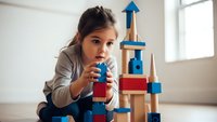 Young girl playing with colorful wooden building blocks creating tower structure in bright playroom developing creativity and motor skills.