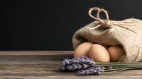 Fresh farm eggs in rustic burlap sack with lavender sprigs on wooden table against dark background, representing organic food and country lifestyle.