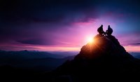 Silhouettes of two climbers resting on mountain peak at sunset with dramatic purple sky and sunburst, mountainous landscape stretches into horizon.