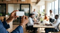 Man holding blank white business card in modern office environment with diverse colleagues working in background.