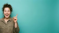 Young woman with brown hair and fair complexion wearing khaki shirt pointing upward with excited expression against turquoise background for advertising and promotional campaigns.