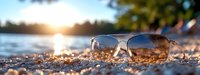 Stylish sunglasses resting on sandy beach at golden hour sunset with ocean waves and tropical palm trees creating perfect summer vacation atmosphere.