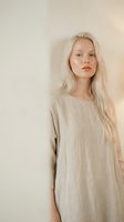 Young blonde Caucasian woman in beige linen dress standing against neutral wall background for fashion and lifestyle photography projects.