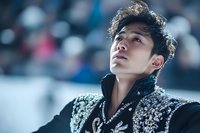 Young Asian male athlete in ornate black performance costume gazes upward with determined expression against blurred arena background, dramatic lighting.
