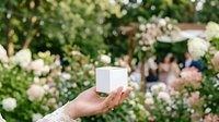 Caucasian woman's hand holding blank white product box in blooming garden with pink and white flowers, ideal for cosmetics, skincare, or gift packaging mockups.