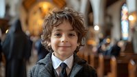 Young boy with curly brown hair wearing formal suit and tie smiling in church interior with stained glass windows and warm lighting, celebrating special occasion.