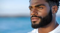 Portrait of confident African American man with well-groomed beard and earring against blurred ocean background, wearing white sweater, looking into distance.