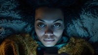 Young person with curly hair illuminated by phone screen in dark room, wrapped in fur blanket, creating moody atmospheric lighting effect.