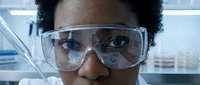 Female scientist of African descent wearing protective safety goggles in laboratory setting, conducting research and analysis work in modern scientific facility.