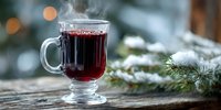 Steaming glass of mulled wine on rustic wooden surface with snowy pine branches and bokeh lights in background for winter holiday celebrations.