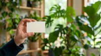 Person holding blank white business card in hand with lush green plants and wooden shelving in background, suitable for branding, corporate identity, and botanical business concepts.