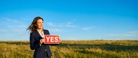Woman in business attire holding yes sign in open field under blue sky, representing positive decision, agreement, approval, success, and affirmative response for corporate concepts.