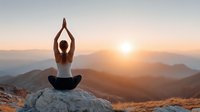 Woman practicing yoga meditation pose on mountain peak during golden sunrise with peaceful valley landscape below.