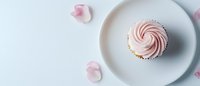 Pink swirled frosting tops delicate cupcake on minimal white ceramic plate, scattered rose petals create romantic mood against light background.