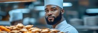 Male baker with dark skin tone and beard wearing light blue chef's hat and uniform in commercial kitchen with fresh baked goods, suitable for bakery, food service, culinary training and restaurant industry marketing.