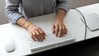 Professional woman typing on white keyboard at modern office desk wearing gray ribbed sweater with hands positioned over computer workspace.