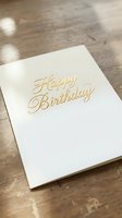Elegant white birthday card with gold foil lettering on rustic wooden table surface for celebration and greeting card design projects.