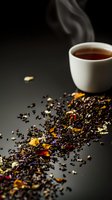 Steaming cup of aromatic tea with scattered loose leaf black tea blend decorated with colorful dried flower petals and herbs on dark background.