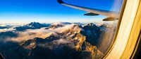 Aerial view from airplane window showing snow-capped mountain peaks bathed in golden sunrise light with clouds below, perfect for travel and aviation content.