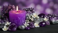 Purple candle burning surrounded by amethyst crystals, lavender sprigs and white flowers on dark surface for spa relaxation and meditation wellness.