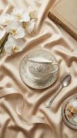 Elegant tea cup with saucer on luxurious silk fabric alongside white orchids and vintage book creating sophisticated afternoon tea setting in warm beige tones.