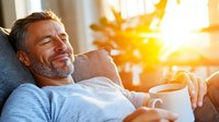 Mature Caucasian man relaxing on couch with coffee cup at sunrise, enjoying peaceful morning moment in casual home setting with golden sunlight through window.
