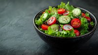 Fresh vegetable salad with cherry tomatoes, cucumber slices, and green lettuce in black ceramic bowl on dark background. Healthy eating concept.