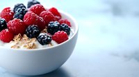 Fresh raspberries and blackberries on top of creamy yogurt with granola in white ceramic bowl against light marble background, healthy breakfast concept.