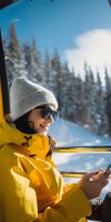 Young woman in yellow ski jacket and beanie using smartphone in cable car with snowy mountain forest view through window during winter vacation.
