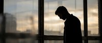 Silhouette of contemplative man standing by large windows in modern office building during sunset with urban cityscape background for business concepts.