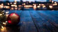 Red and gold Christmas ornament on dark wooden surface with glowing string lights and blurred city bokeh background creates festive holiday atmosphere.