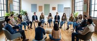 Diverse group of professionals sitting in circle during corporate team building meeting or support group session in modern office space with wooden floor and natural light.
