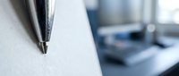 Ballpoint pen tip on white paper with blurred office workspace background, suitable for business writing, education, and stationery concepts.