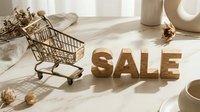 Wooden SALE letters and miniature shopping cart on marble surface with dried flowers, soft natural light, retail discount concept.