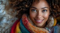 Young African American woman with natural curly hair wearing colorful knitted winter scarf, showing bright genuine smile and blue eyes in warm atmospheric lighting.