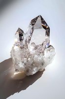 Clear quartz crystal cluster with multiple points catching light and casting shadows, showcasing natural geometric formation and prismatic reflections on gray background.