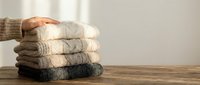 Hand protecting stack of folded knit sweaters in neutral tones on wooden surface. Cozy winter clothing care and storage concept for seasonal wardrobe organization.