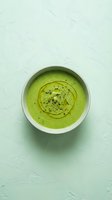 Creamy green soup with olive oil swirl and black pepper in white bowl on textured mint background for healthy cooking and restaurant menu design.