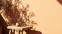 Warm sunlight creates dramatic plant shadows on textured wall with wooden stool and ceramic cup, perfect for minimalist interior design and lifestyle content.