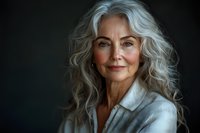 Elegant mature woman with long wavy silver hair and natural makeup wearing white blazer smiles confidently against dark background, representing aging gracefully.