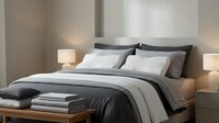 Contemporary bedroom with neatly made bed featuring gray and white bedding, decorative pillows, folded linens on bench, and warm ambient lighting from bedside lamps.