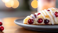 Fresh cranberry scone with white icing drizzle on gray plate against blurred bokeh background, close-up view showing moist texture and bright red berries.