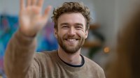 Friendly young Caucasian man with curly hair and beard waving hello with warm smile in casual sweater. Greeting gesture for social media and communication.