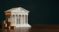 Classical bank building model with golden coins stacks on wooden surface against dark background representing financial services and monetary institutions.