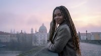 Young African American woman with dreadlocks smiling at sunset in urban European city square wearing cozy sweater for lifestyle and travel content.