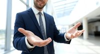 Professional businessman in navy suit with open hands presenting concept in modern office lobby with natural lighting for corporate presentations.