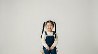 Young Asian girl with braided pigtails wearing denim overalls and cream sweater, smiling at camera against neutral background for portrait and lifestyle use.