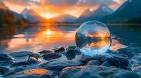 Crystal sphere on rocky shore reflecting mountain landscape at sunset with vibrant orange sky over tranquil lake waters.