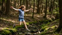 Young girl jumping over forest stream with arms spread wide showing joy and freedom in natural woodland setting for outdoor adventure concepts.